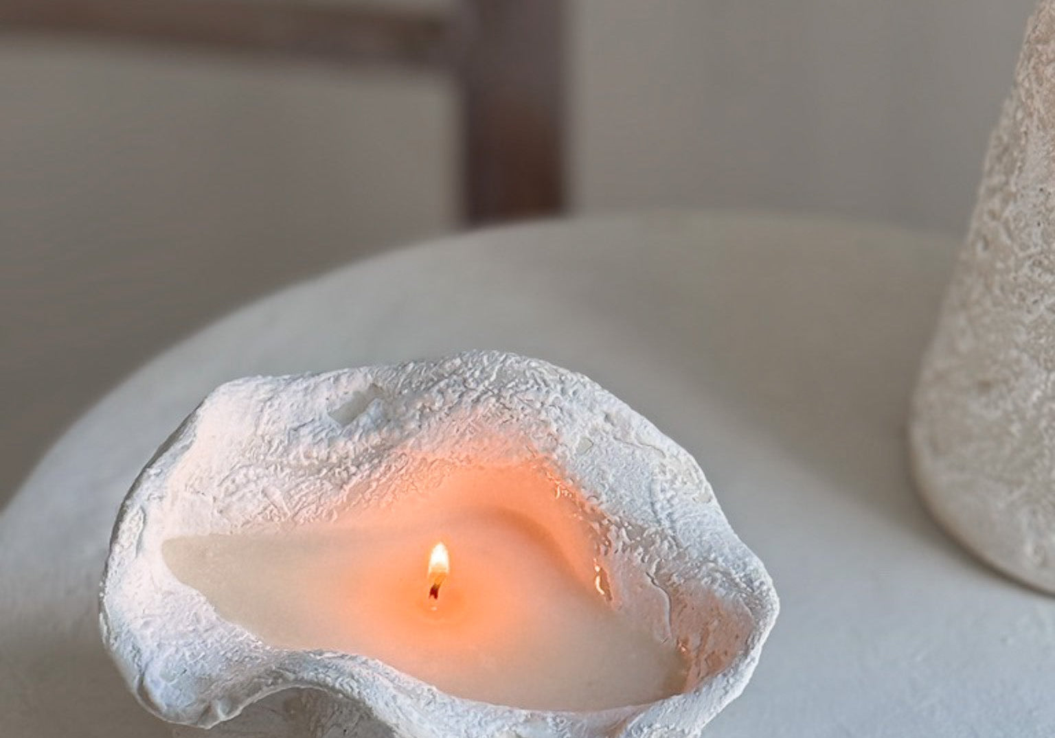 A handmade ruffled edge ceramic vessel candle burning on a white pedestal table, its wax pool glowing amber, a rustic wooden beam behind