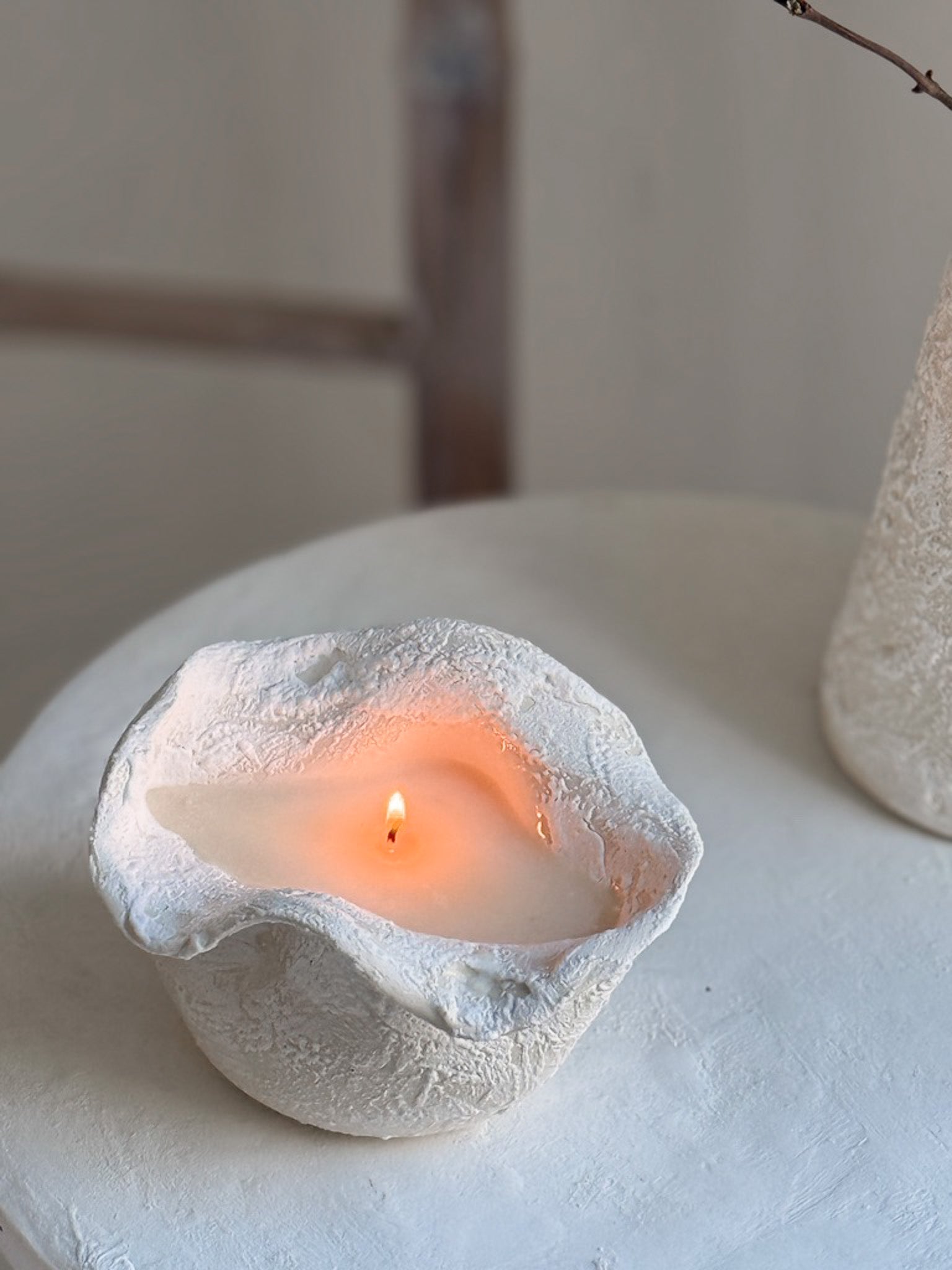 A handmade ruffled edge ceramic vessel candle burning on a white pedestal table, its wax pool glowing amber, a rustic wooden beam behind