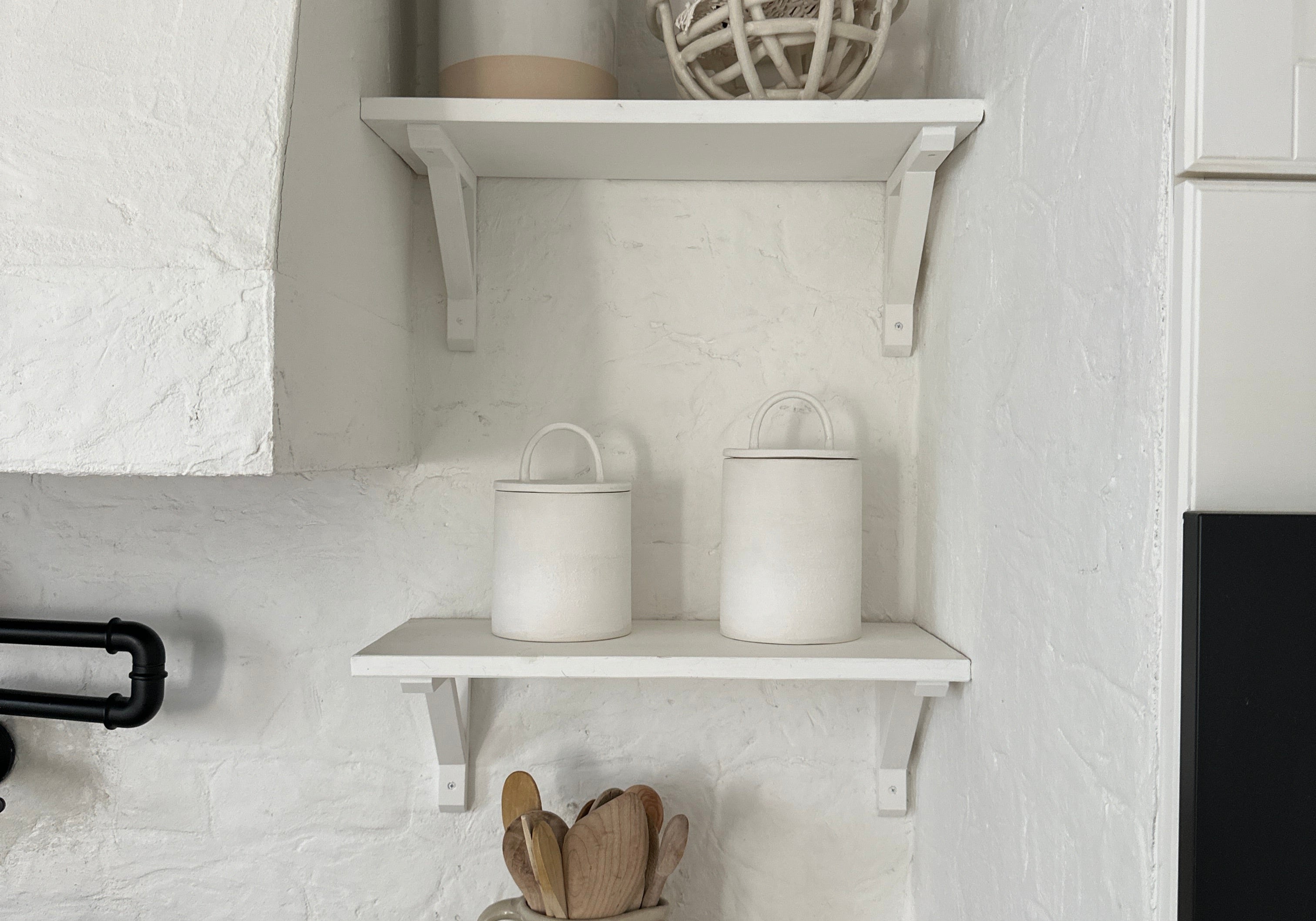 Two handmade ceramic canisters with long-arch lids on a shelf above the stove in a soft, neutral kitchen, with wooden spoons in a ceramic vase