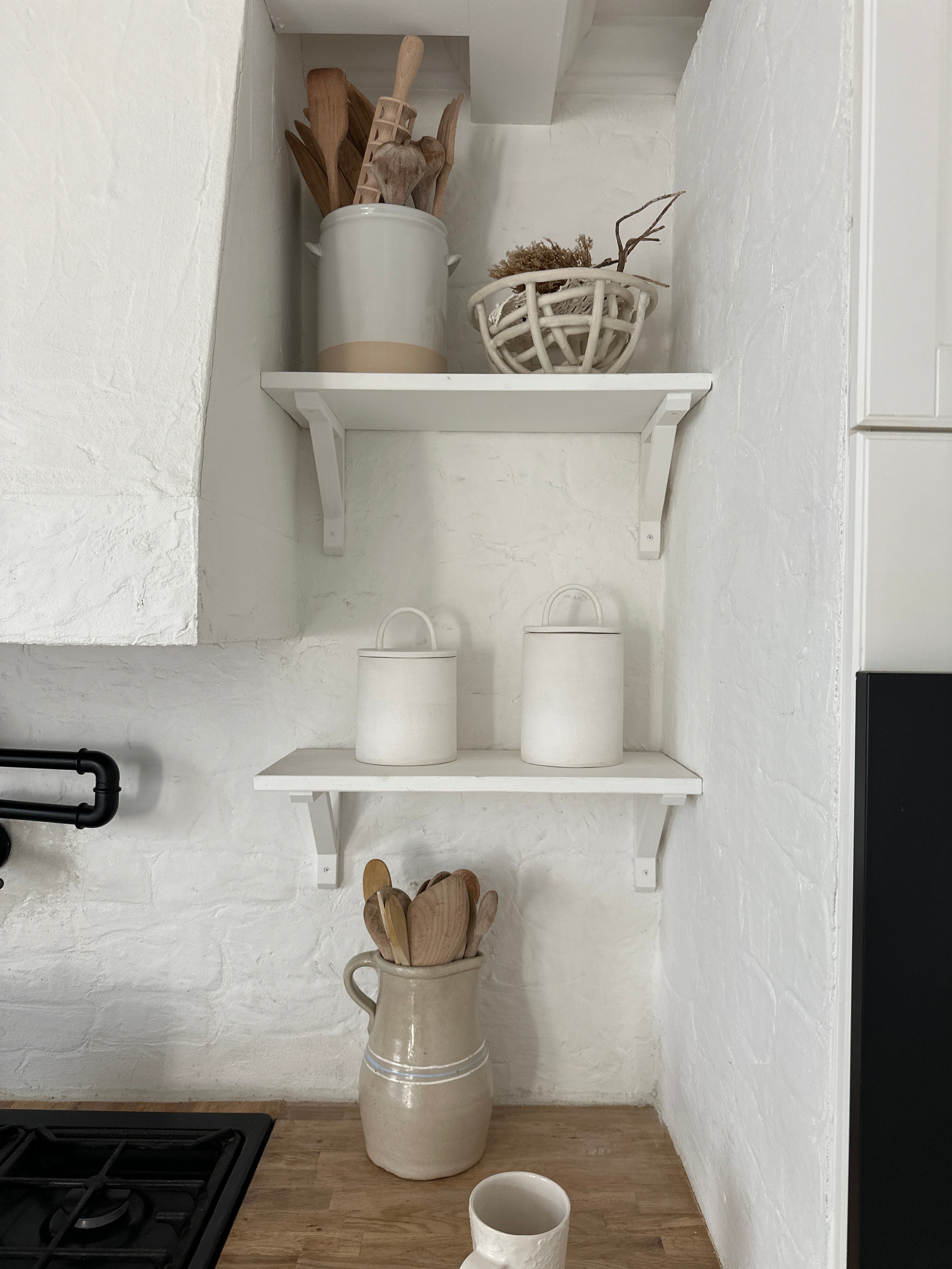 Two handmade ceramic canisters with long-arch lids on a shelf above the stove in a soft, neutral kitchen, with wooden spoons in a ceramic vase