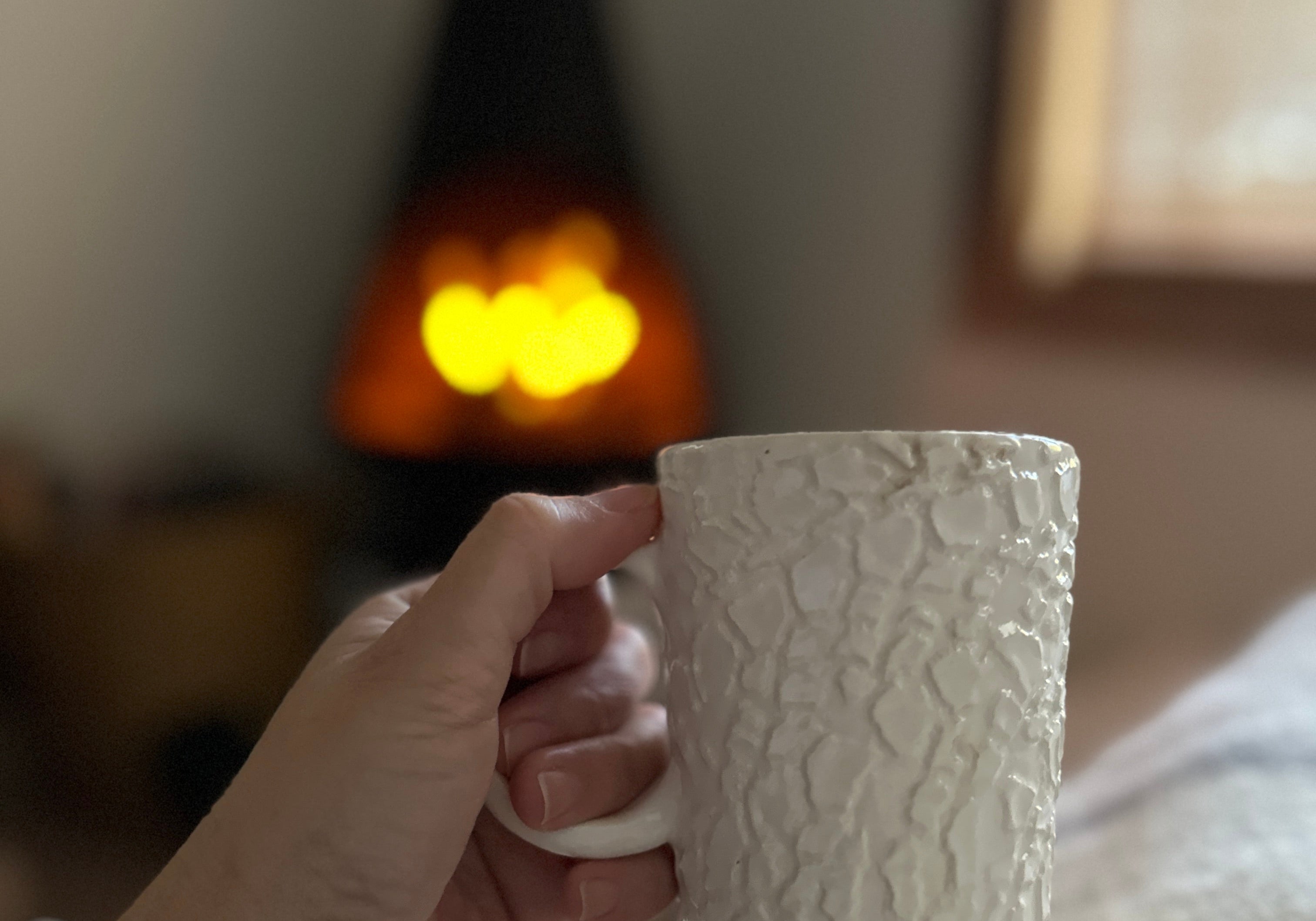 A hand holding a tall textured white ceramic mug, with a lit wood stove and a bright window softly out of focus behind.