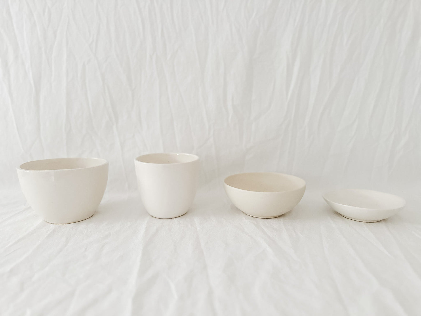 Four gloss white handmade ceramic bowls in a row on rumpled white linen, two deep cups beside two shallow serving bowls, from the annual sample sale