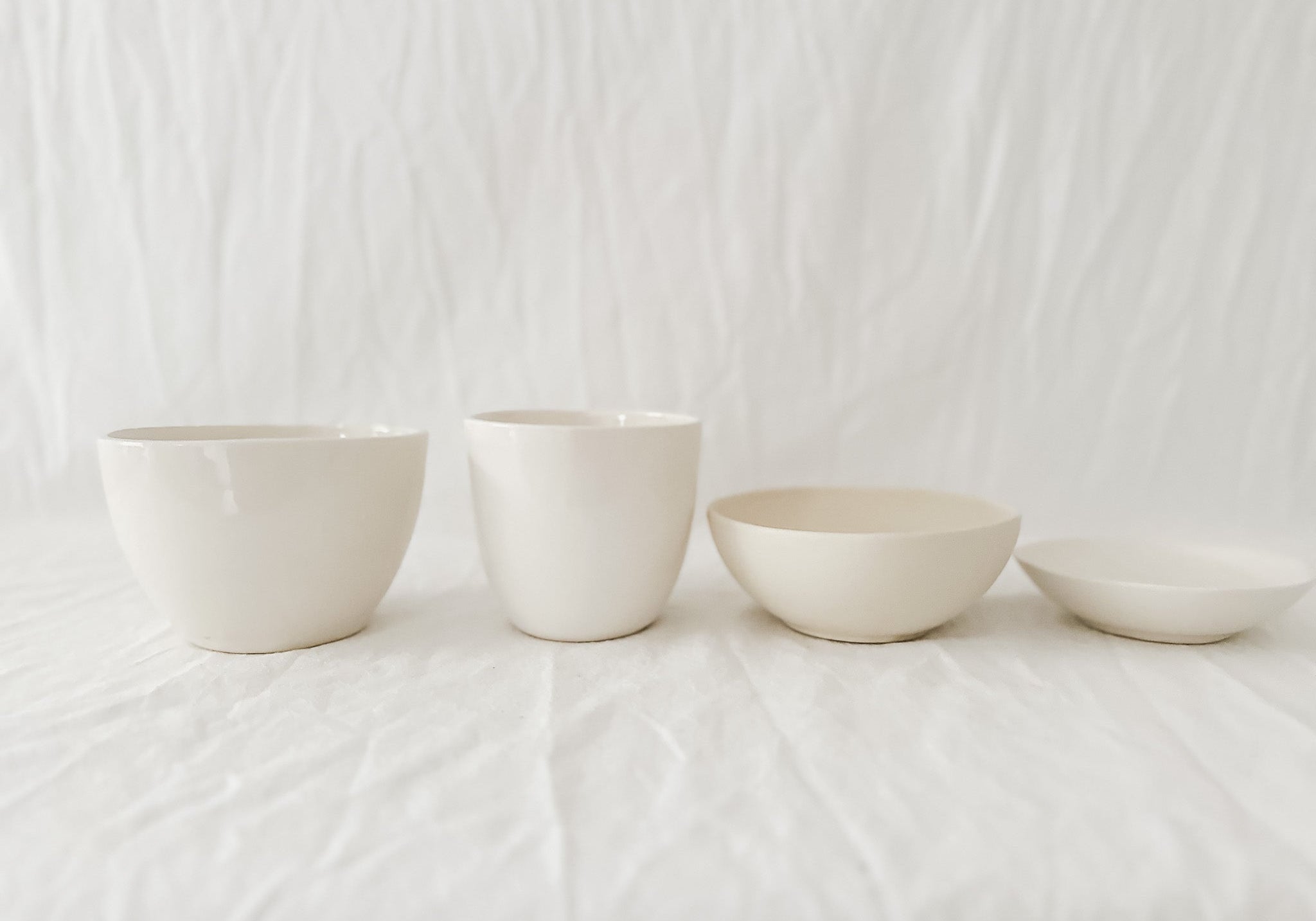 Two straight-sided cream cups and two shallow handmade stoneware bowls in a row on rumpled white linen against a soft white curtain