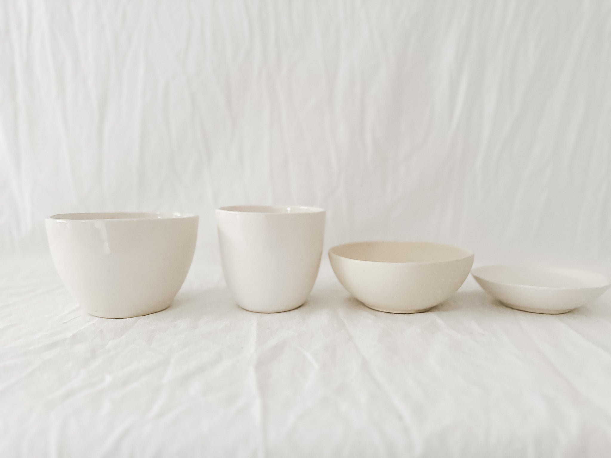 Two straight-sided cream cups and two shallow handmade stoneware bowls in a row on rumpled white linen against a soft white curtain