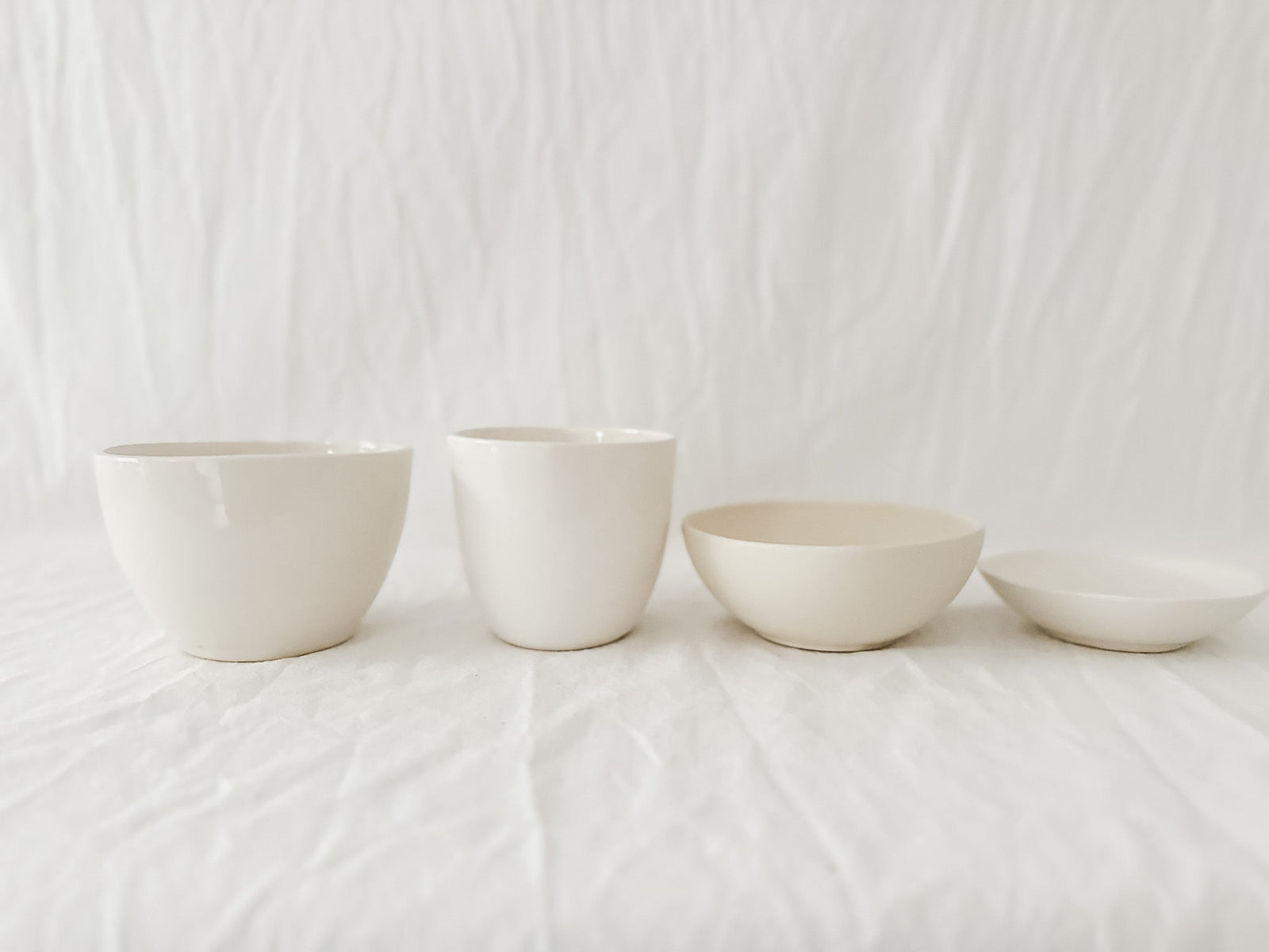 Two straight-sided cream cups and two shallow handmade stoneware bowls in a row on rumpled white linen against a soft white curtain