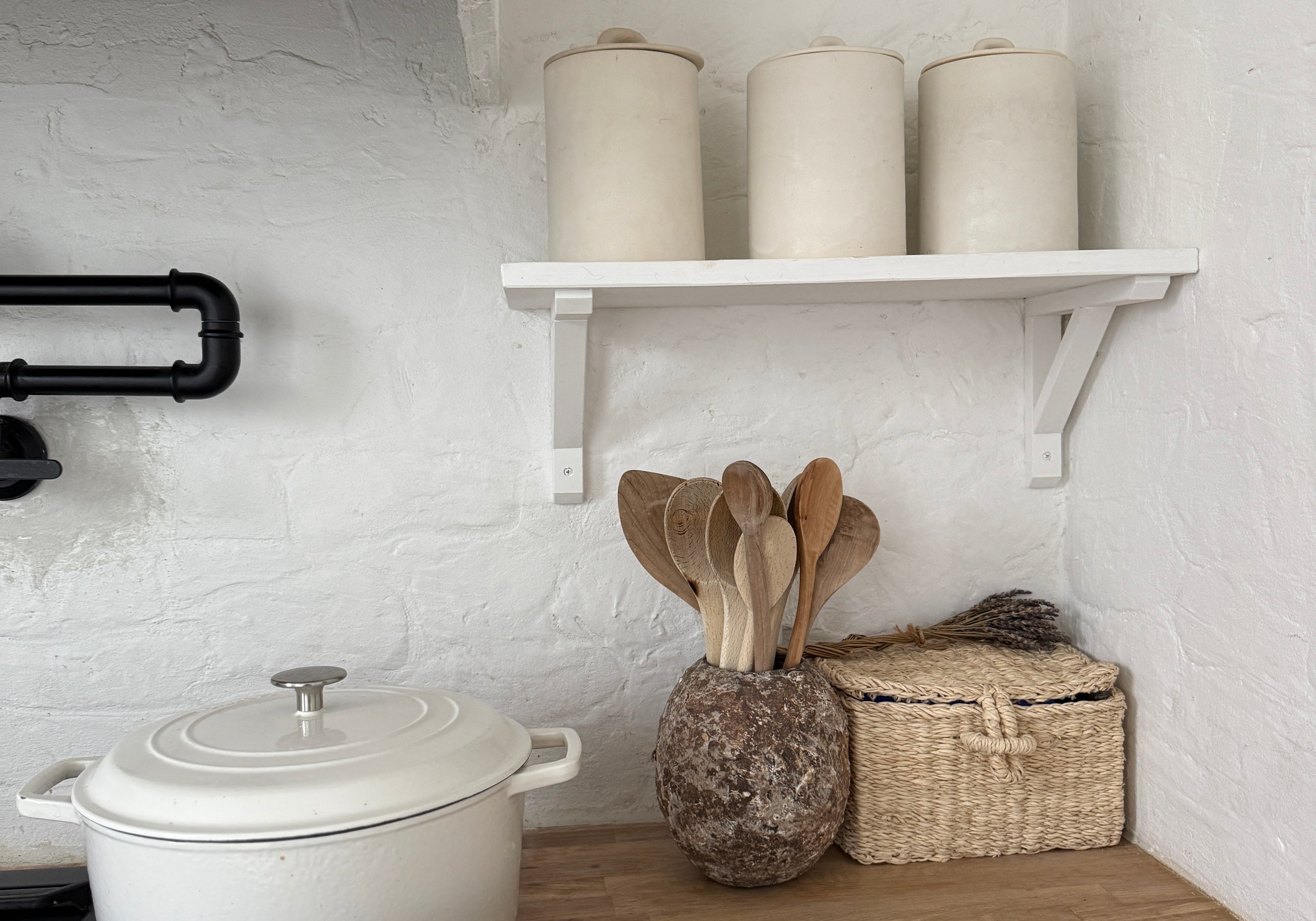 A white kitchen corner with a cream enamel pot on the hob, three cream ceramic canisters on an open shelf, and a mottled stoneware crock of wooden spoons beside a woven basket on a clear wood counter