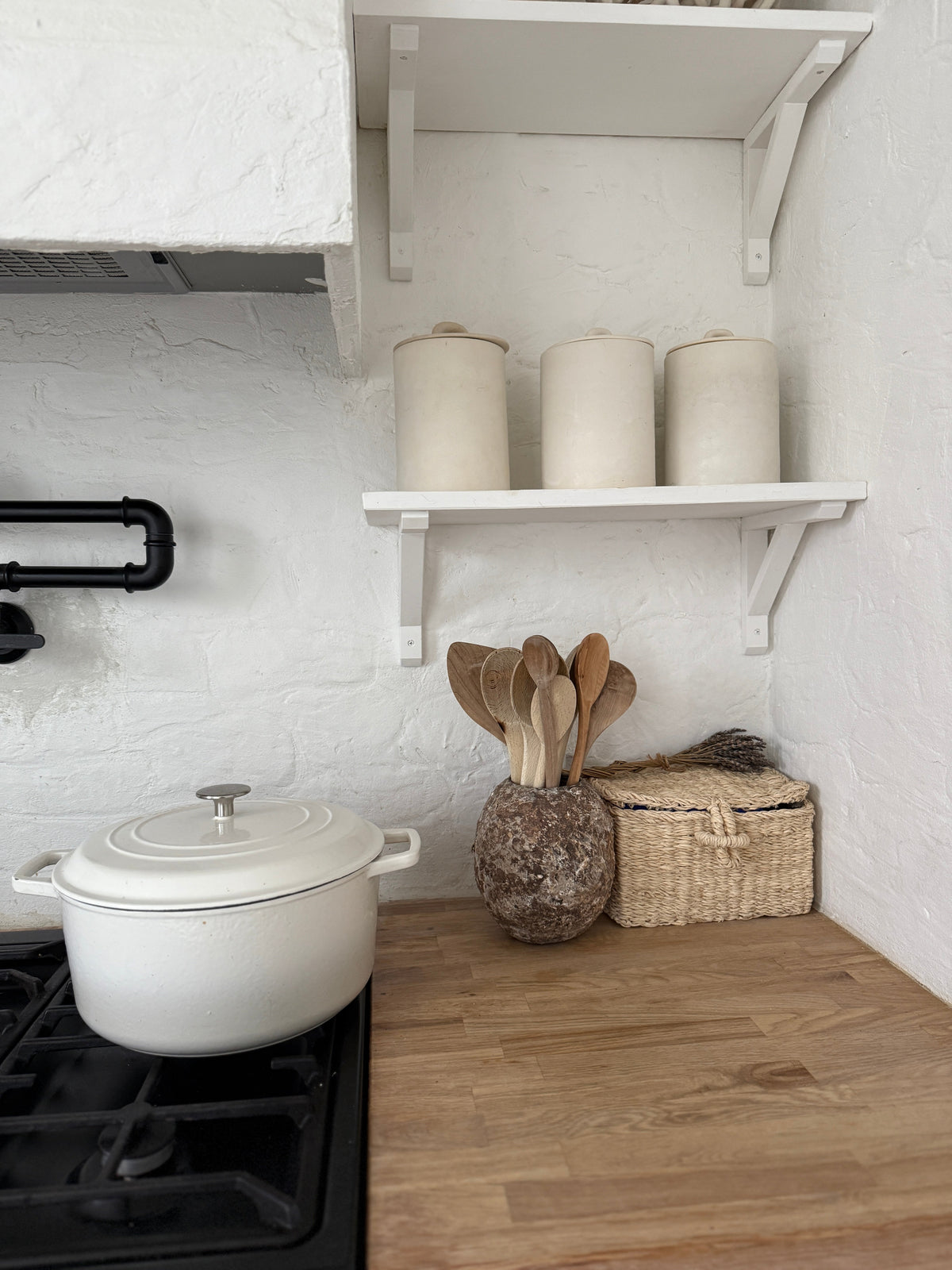 A white kitchen corner with a cream enamel pot on the hob, three cream ceramic canisters on an open shelf, and a mottled stoneware crock of wooden spoons beside a woven basket on a clear wood counter