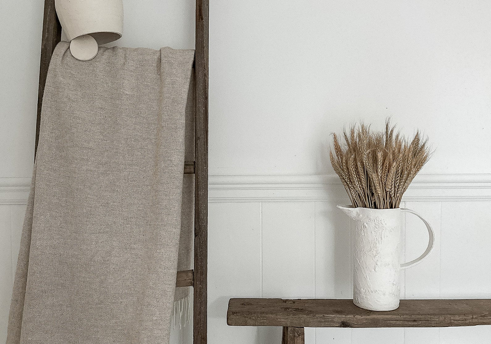 Quiet sunlit summer corner with pale pottery, natural wood, and dried stems.