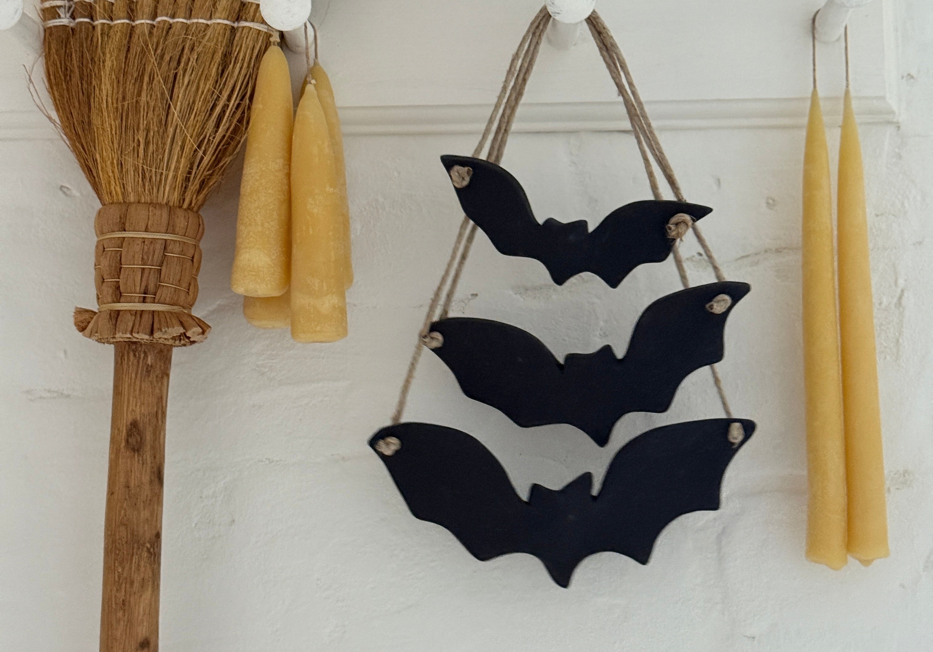 Three black stoneware bats strung on jute rope hanging on a white plaster wall between two beeswax taper candles, a rustic broom beside them