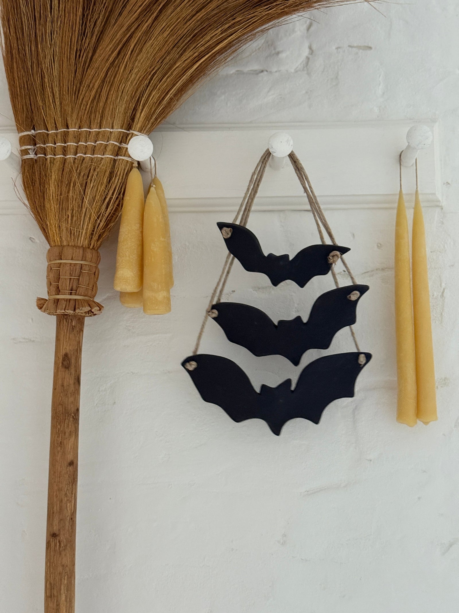 Three black stoneware bats strung on jute rope hanging on a white plaster wall between two beeswax taper candles, a rustic broom beside them