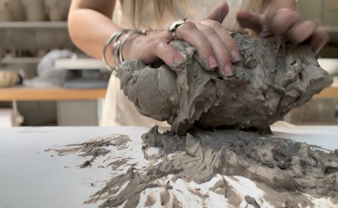 Two hands stacked with silver rings pressing into a large lump of raw grey stoneware clay on a white studio worktable, shelves behind