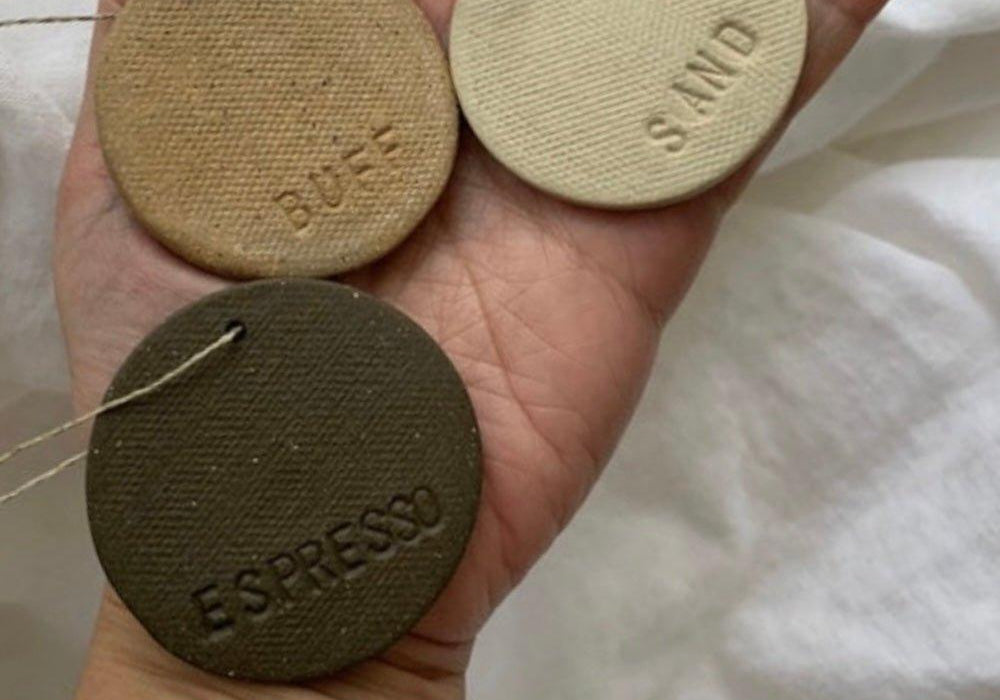 An open palm holding three fired ceramic glaze test discs incised Buff, Sand and Espresso, threaded on a jute cord against white linen