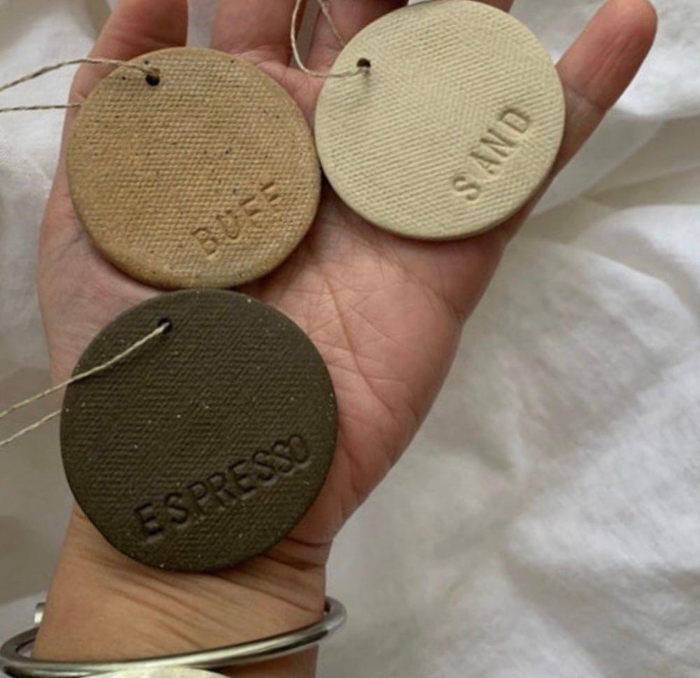An open palm holding three fired ceramic glaze test discs incised Buff, Sand and Espresso, threaded on a jute cord against white linen