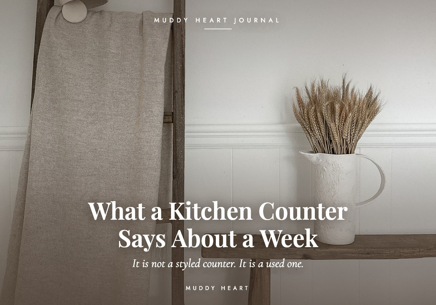 A white handmade ceramic pitcher of dried wheat on a dark wooden bench beside a linen towel hung on a ladder, titled 'What a Kitchen Counter Says About a Week' — Muddy Heart Journal