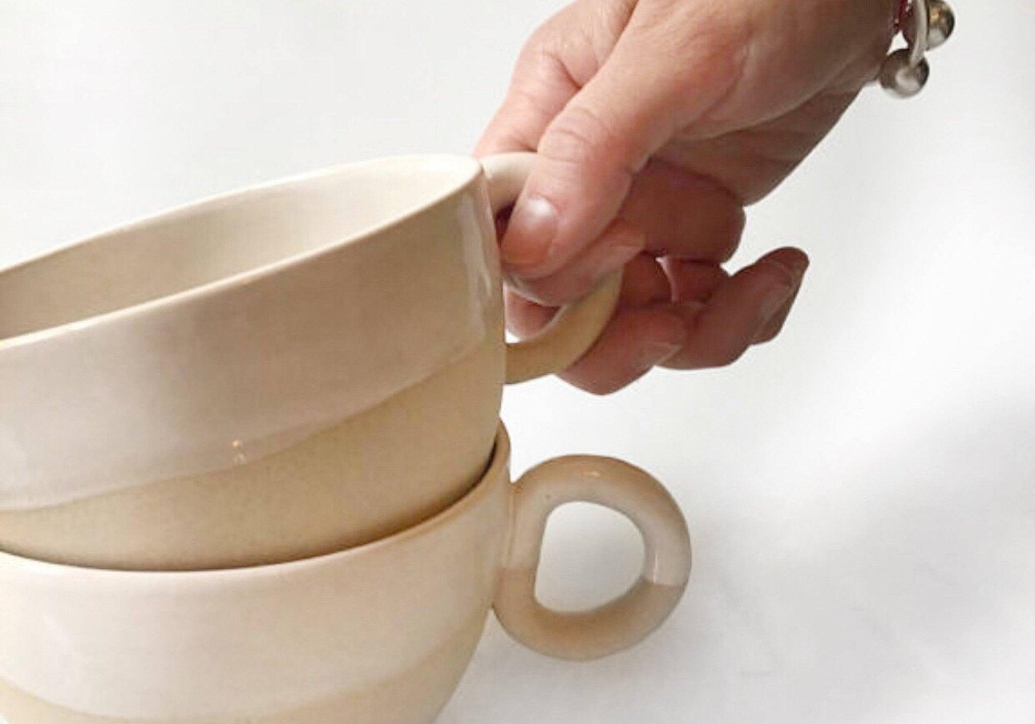 A hand lifting the top mug from two stacked Natural Sand handmade ceramic mugs, thumb hooked through the thick round ring handle