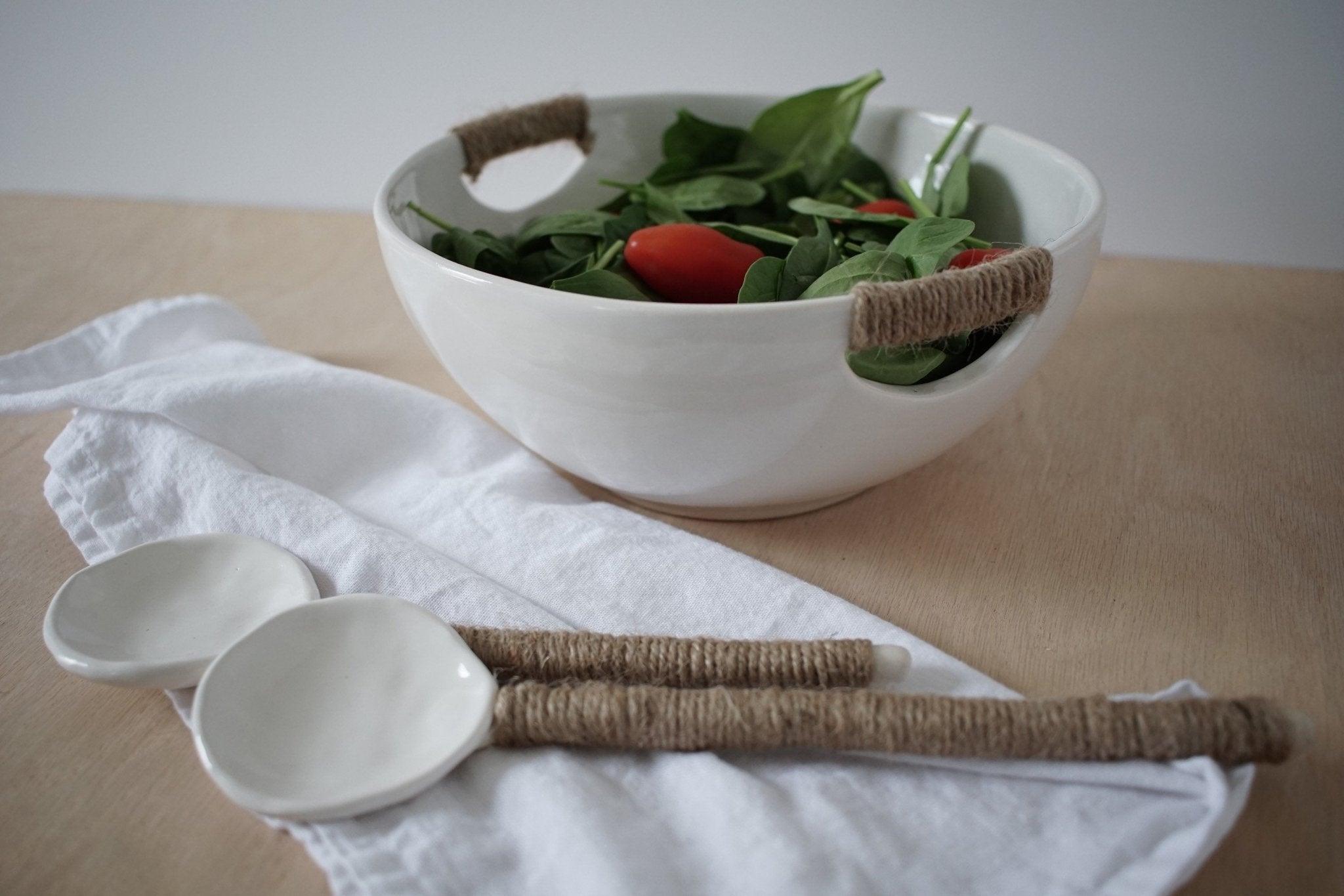 A white stoneware serving bowl with jute-wrapped rope handles holding a green salad and cherry tomatoes, two ceramic serving spoons on linen beside it