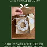 The image is a promotional poster for Christmas delivery orders. It features a hand placing a white ornament into a wooden box, which is placed on a wooden surface. The text on the poster indicates that orders are placed by December 5th and that the ship will be in time for Christmas delivery. The text also mentions that orders received after December 5th will be shipped in January.